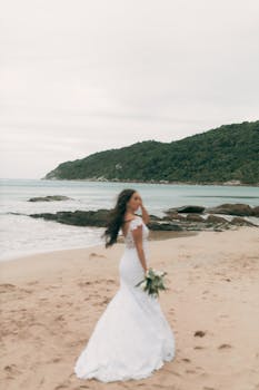 A bride in a white dress poses elegantly by a tranquil beach with lush hills in the background.
