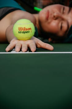A tennis player rests on a court with a Wilson ball in focus, conveying exhaustion.