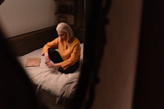 An elderly woman with white hair sits alone on a bed, wearing a yellow top. The scene is introspective and intimate.