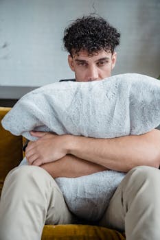 Man sitting indoors with a pillow, showing signs of emotional stress and contemplation.