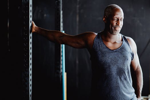 Middle-aged man stretching arm in gym setting while wearing a gray tank top. Fitness and wellness concept.