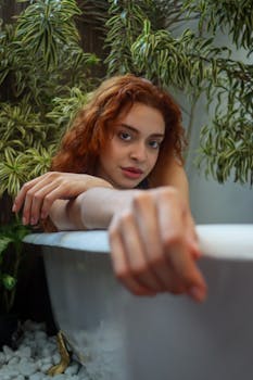 Red-haired woman in a bathtub surrounded by lush plants, evoking relaxation and nature.